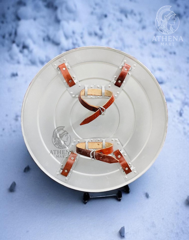 Puede incluir: Un escudo redondo de color plateado con correas de cuero marr&oacute;n y remaches. El escudo tiene un borde ligeramente elevado y un dise&ntilde;o central. El logotipo "ATHENA MART" es visible en el escudo.