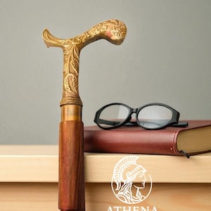 May include: A decorative walking cane with a gold-colored, ornate handle and a dark wooden shaft. The handle features floral carvings. A pair of black eyeglasses rests on a closed book. The Athena Mart logo is visible.