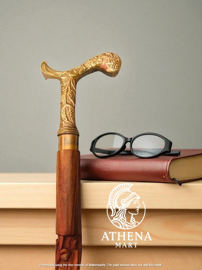 May include: A decorative walking cane with a gold-colored, ornate handle and a dark wooden shaft. The handle features floral carvings. A pair of black eyeglasses rests on a closed book. The Athena Mart logo is visible.