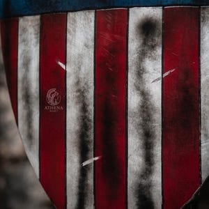 May include: Close-up of a shield with a distressed American flag design. The shield features alternating red and white stripes with a blue top. The shield has a weathered appearance, with scratches and dark areas.