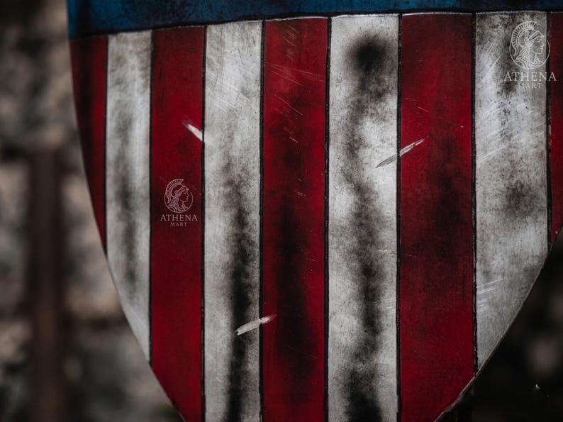 May include: Close-up of a shield with a distressed American flag design. The shield features alternating red and white stripes with a blue top. The shield has a weathered appearance, with scratches and dark areas.