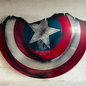 May include: A distressed Captain America shield with a red, white, and blue color scheme. The shield features a central blue circle with a white star, surrounded by red and white stripes. The shield has a worn, battle-damaged appearance.