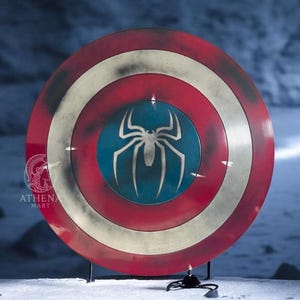 Puede incluir: Un escudo redondo con un dise&ntilde;o rojo, blanco y azul. El escudo presenta un gran emblema de ara&ntilde;a blanca sobre un centro azul. El escudo est&aacute; sostenido por un soporte negro. El escudo es un accesorio o art&iacute;culo de colecci&oacute;n.