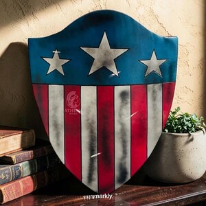 May include: A shield with a blue top section featuring three white stars, transitioning to red and white stripes. The shield has a distressed, vintage appearance. The Athena Mart logo is visible on the shield. A stack of old books and a potted plant are in the background.