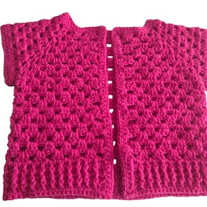 May include: A bright fuchsia crochet cardigan featuring a classic granny square stitch pattern throughout the body and short sleeves. The garment has an open front design with a ribbed hem and neckline, crafted from a vibrant pink yarn material.
