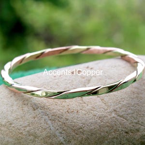 May include: A twisted metal bangle bracelet with copper and gold hues. The bracelet is displayed on a light-coloured stone. The text "Accents1Copper" is visible on the stone.