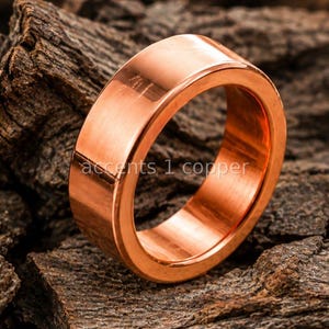 May include: A polished copper ring with a smooth, flat outer surface and a rounded interior. The metal has a warm, reddish-orange metallic luster. The text "accents 1 copper" is visible on the surface of the ring, reflecting the light.