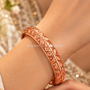Può includere: Un bracciale rigido in metallo color rame caratterizzato da un intricato motivo in rilievo con forme geometriche ripetitive e linee curve. La superficie del bracciale ha una finitura lucida che riflette la luce, mettendo in risalto le incisioni.