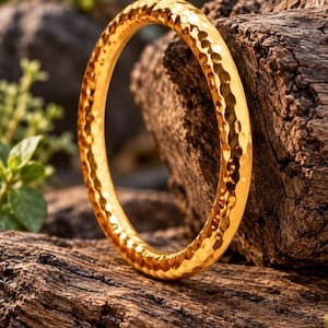 May include: A circular gold-coloured bangle bracelet featuring a hammered, textured surface that reflects light. The bangle has a rounded profile and a polished finish, displaying a consistent metallic sheen across its entire circumference.