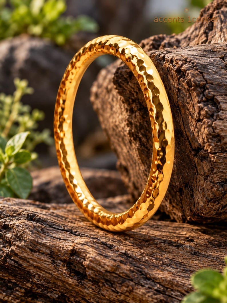 May include: A circular gold-coloured bangle bracelet featuring a hammered, textured surface that reflects light. The bangle has a rounded profile and a polished finish, displaying a consistent metallic sheen across its entire circumference.