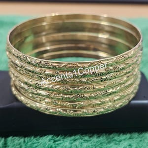 Puede incluir: Un conjunto de pulseras doradas con un dise&ntilde;o floral en relieve. Las pulseras est&aacute;n apiladas una encima de la otra, y el texto "Accents 1 Copper" es visible en el lateral de las pulseras.