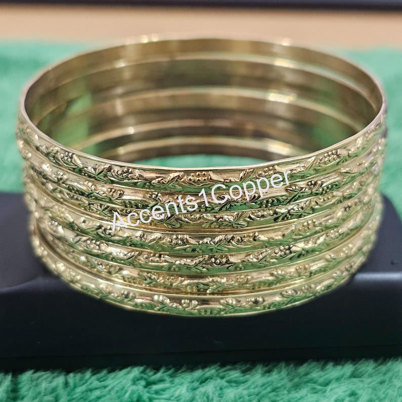 Puede incluir: Un conjunto de pulseras doradas con un dise&ntilde;o floral en relieve. Las pulseras est&aacute;n apiladas una encima de la otra, y el texto "Accents 1 Copper" es visible en el lateral de las pulseras.