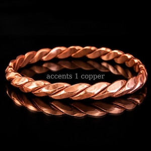 May include: A polished copper bangle bracelet featuring a continuous twisted rope design. The metallic surface has a warm, reddish-brown hue with reflective highlights. The text "accents 1 copper" is printed in the center of the circular bangle.