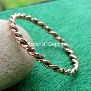 May include: A copper-colored twisted bangle bracelet. The bracelet has a spiral design and is resting on a stone. The background is a blurred green. The text "Accents1Copper" is visible on the stone.