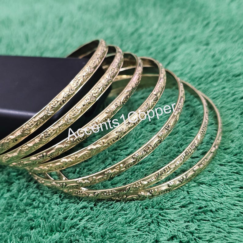 Puede incluir: Un conjunto de pulseras doradas con un dise&ntilde;o floral en relieve. Las pulseras est&aacute;n apiladas sobre una superficie negra, sobre un fondo verde. El texto "Accents1Copper" es visible en las pulseras.