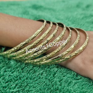 Puede incluir: Un conjunto de pulseras doradas con un intrincado dise&ntilde;o floral. Las pulseras est&aacute;n apiladas en una mu&ntilde;eca, mostrando sus patrones detallados. El texto "Accents1Copper" es visible en las pulseras.