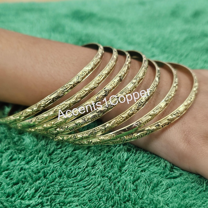 Puede incluir: Un conjunto de pulseras doradas con un intrincado dise&ntilde;o floral. Las pulseras est&aacute;n apiladas en una mu&ntilde;eca, mostrando sus patrones detallados. El texto "Accents1Copper" es visible en las pulseras.