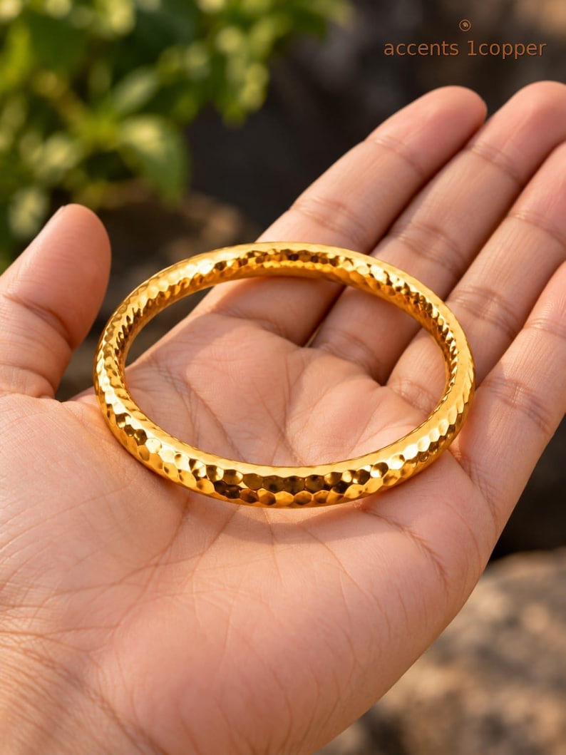 May include: A circular bangle bracelet with a bright gold-tone finish and a hammered, textured surface. The metal band features a repeating indented pattern across its entire exterior, creating a reflective appearance. The text accents 1copper is visible.