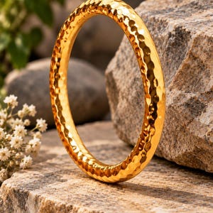 May include: A circular gold-coloured bangle bracelet featuring a hammered, dimpled texture across its entire surface. The metal has a reflective, polished finish that catches the light, creating a distinct pattern of small, concave indentations.