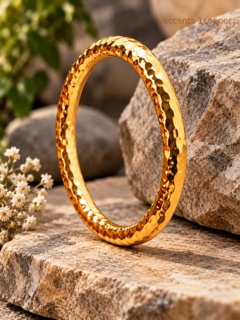 May include: A circular gold-coloured bangle bracelet featuring a hammered, dimpled texture across its entire surface. The metal has a reflective, polished finish that catches the light, creating a distinct pattern of small, concave indentations.