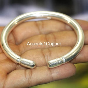 Może przedstawiać: Błyszcząca, srebrna bransoletka typu mankiet. Otwarta konstrukcja ma zaokrąglone zakończenia. Tekst "Accents1Copper" jest widoczny na zdjęciu. Ta bransoletka to biżuteria.