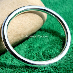 May include: A shiny, silver-colored bangle bracelet. The circular bracelet is displayed on a green, textured surface. The text "Accents1Copper" is visible on the background.