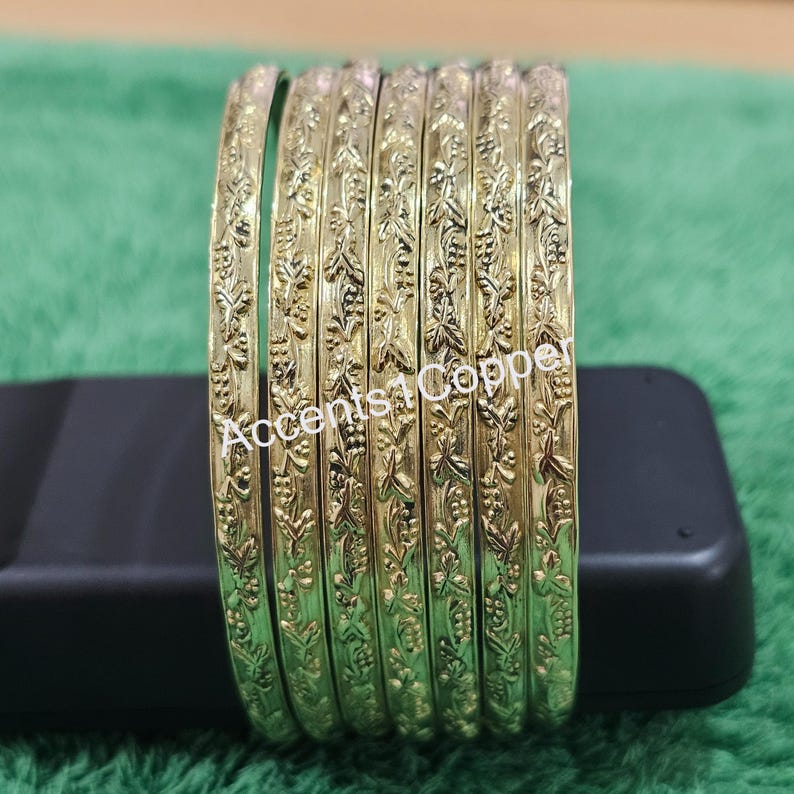 Puede incluir: Un conjunto de pulseras doradas con un dise&ntilde;o floral en relieve. Las pulseras est&aacute;n sobre una superficie negra, sobre un fondo verde. El texto "Accents1Copper" es visible en las pulseras.