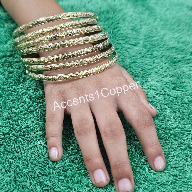 Puede incluir: Brazaletes dorados con un dise&ntilde;o floral en relieve apilados en una mano. Los brazaletes se muestran sobre una superficie verde. El texto "Accents1Copper" es visible en la mano.