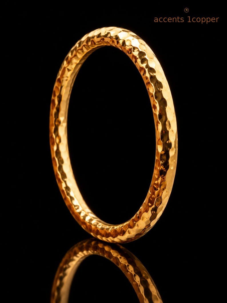 May include: A circular bangle bracelet with a metallic copper finish and a hammered, textured surface. The jewellery piece features a reflective, uneven exterior that catches light. The text 'accents 1copper' is visible in the upper right corner.