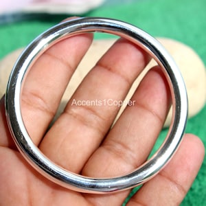 May include: A polished, silver-coloured bangle bracelet. The smooth, rounded bracelet is held in a person's hand. The background is a light tan colour. The bracelet is a simple, elegant design.