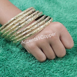 Puede incluir: Un conjunto de pulseras doradas con un dise&ntilde;o floral en relieve. Las pulseras est&aacute;n apiladas en una mano, con el texto "Accents1Copper" visible. Las pulseras est&aacute;n sobre un fondo verde.