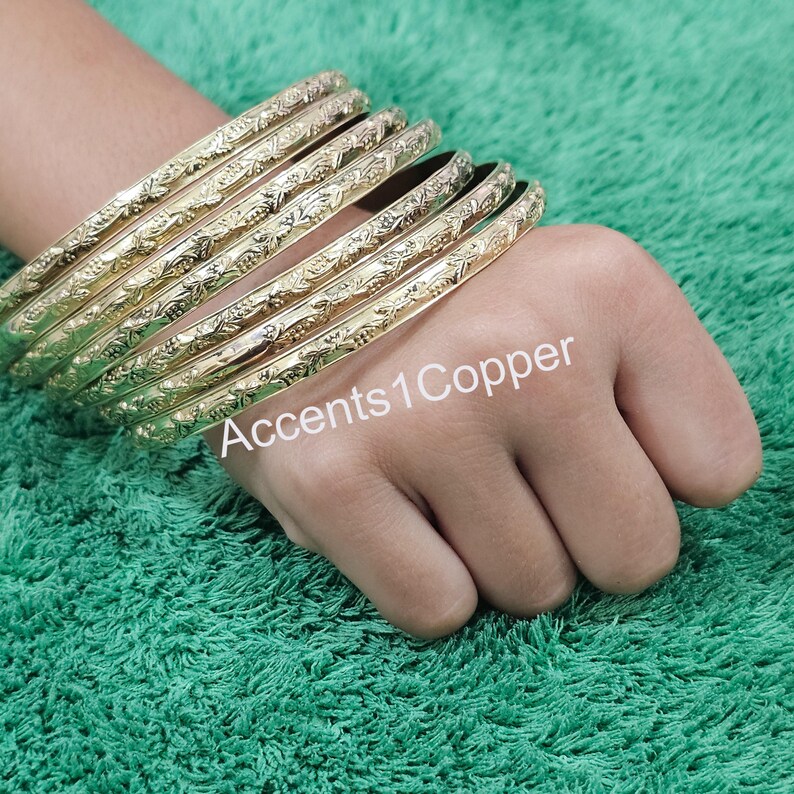 Puede incluir: Un conjunto de pulseras doradas con un dise&ntilde;o floral en relieve. Las pulseras est&aacute;n apiladas en una mano, con el texto "Accents1Copper" visible. Las pulseras est&aacute;n sobre un fondo verde.