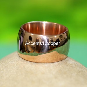 Può includere: Un anello largo in rame lucido dalla forma arrotondata. L'anello ha una superficie lucida e riflettente e il testo "Accents1Copper" è visibile sull'anello. L'anello è appoggiato su una superficie marrone chiaro con uno sfondo verde.