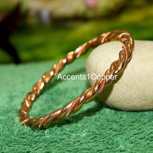 May include: A copper-colored twisted bangle bracelet. The bracelet has a braided design with a reddish-brown hue. The text "Accents 1 Copper" is visible on the image. The bracelet is resting on a green surface.