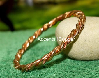 Puede incluir: Una pulsera de brazalete retorcida de color cobre. La pulsera tiene un diseño trenzado con un tono marrón rojizo. El texto "Accents 1 Copper" es visible en la imagen. La pulsera descansa sobre una superficie verde.
