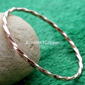 May include: A copper-colored, twisted metal bangle bracelet. The bracelet has a spiral design and is resting on a rock against a green background. The text "Accents1Copper" is visible.