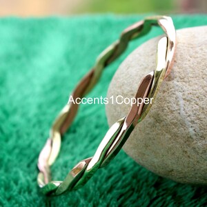 May include: A twisted metal bangle bracelet with a tri-color design of copper, gold, and rose gold. The bracelet is displayed on a green surface with a stone in the background. The text "Accents1Copper" is visible.