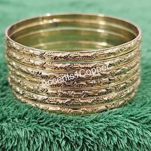 Puede incluir: Una pila de pulseras doradas con un dise&ntilde;o floral en relieve. Las pulseras est&aacute;n apiladas una encima de otra, y el texto "Accents4Copper" es visible en las pulseras.