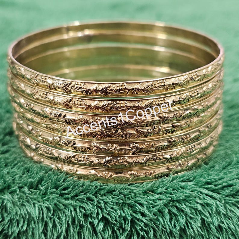 Puede incluir: Una pila de pulseras doradas con un dise&ntilde;o floral en relieve. Las pulseras est&aacute;n apiladas una encima de otra, y el texto "Accents4Copper" es visible en las pulseras.