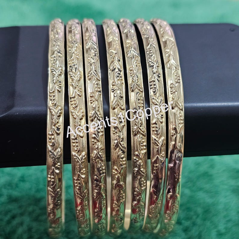 Puede incluir: Un conjunto de siete pulseras doradas con un dise&ntilde;o floral en relieve. Las pulseras est&aacute;n dispuestas una al lado de la otra, mostrando sus intrincados patrones. El texto "Accents4Copper" es visible.