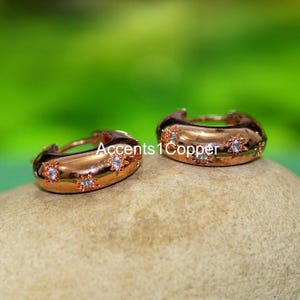 Op de afbeelding: Roségouden oorringen met stervormige accenten en kleine heldere stenen. De oorbellen worden getoond op een lichtbruine steen tegen een vage groene achtergrond. De tekst "Accents 1 Copper" is zichtbaar.