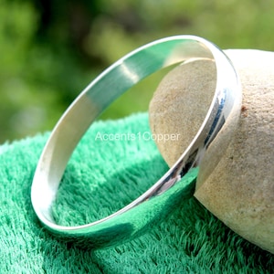 Op de afbeelding: Een glanzende, zilverkleurige armband. De gladde, afgeronde armband wordt getoond op een groene ondergrond en een lichtbruine steen. De armband heeft een eenvoudig, elegant ontwerp en reflecteert het licht. De tekst "Accents1Copper" is zichtbaar.