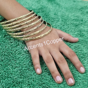 Puede incluir: Un conjunto de pulseras doradas con un dise&ntilde;o floral en relieve. Las pulseras est&aacute;n apiladas unas sobre otras. El texto "Accents1Copper" es visible en la imagen.