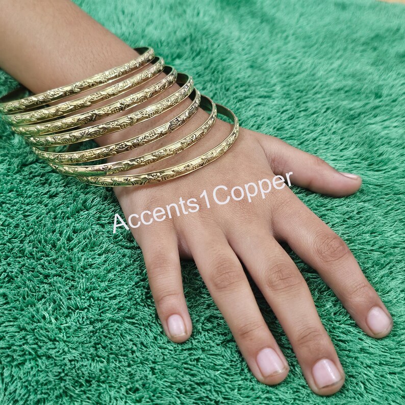 Puede incluir: Un conjunto de pulseras doradas con un dise&ntilde;o floral en relieve. Las pulseras est&aacute;n apiladas unas sobre otras. El texto "Accents1Copper" es visible en la imagen.