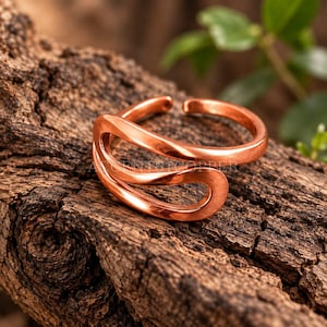Könnte beinhalten: Ein polierter Kupferring mit offenem Bypass-Design, bestehend aus zwei glatten, geschwungenen Bändern, die sich überlappen. Das Metall hat einen warmen, metallisch orange-braunen Farbton und eine reflektierende Oberfläche. Der Text ACCENTSCOPPER ist sichtbar.