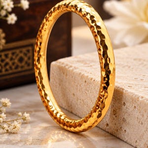 May include: A gold-coloured bangle bracelet featuring a hammered, textured surface that creates a reflective, dimpled appearance. The circular jewellery piece has a polished finish and is displayed upright, showcasing its consistent rounded profile.