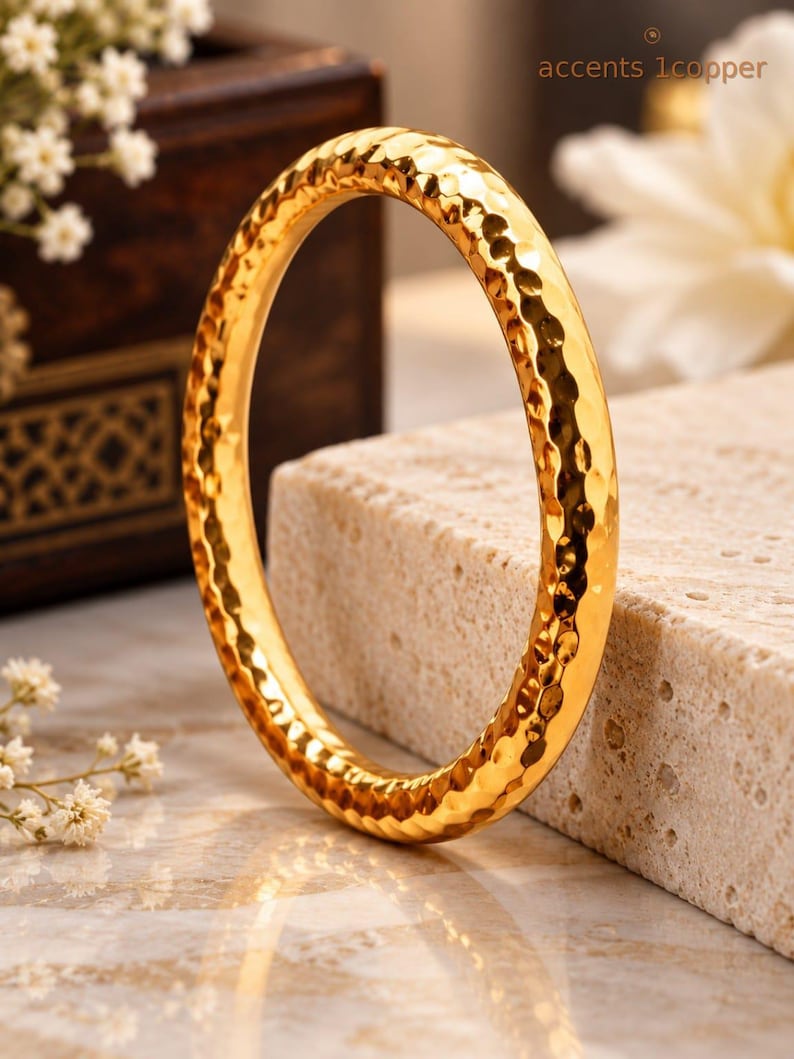 May include: A gold-coloured bangle bracelet featuring a hammered, textured surface that creates a reflective, dimpled appearance. The circular jewellery piece has a polished finish and is displayed upright, showcasing its consistent rounded profile.