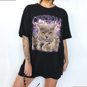 Puede incluir: Camiseta negra con un gr&aacute;fico de un gato gris y gatitos, con la palabra "CUSTOM" arriba. El dise&ntilde;o incluye un rayo p&uacute;rpura y estrellas. La camiseta est&aacute; etiquetada como "Style H".