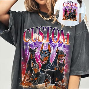 Puede incluir: Camiseta gris oscuro con un gr&aacute;fico de cinco Doberman Pinschers y la palabra "CUSTOM" en degradado. El dise&ntilde;o incluye rayos y el texto "Your Text". Una imagen m&aacute;s peque&ntilde;a del dise&ntilde;o est&aacute; en una camiseta blanca.
