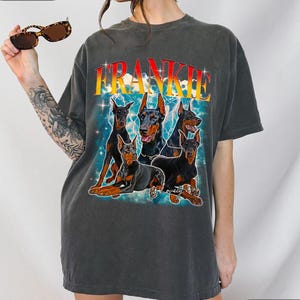 Puede incluir: Camiseta gris oscuro con un gr&aacute;fico de cinco Doberman Pinschers y el nombre "FRANKIE" en una fuente retro. El dise&ntilde;o incluye rayos y la frase "My lovely dogs".
